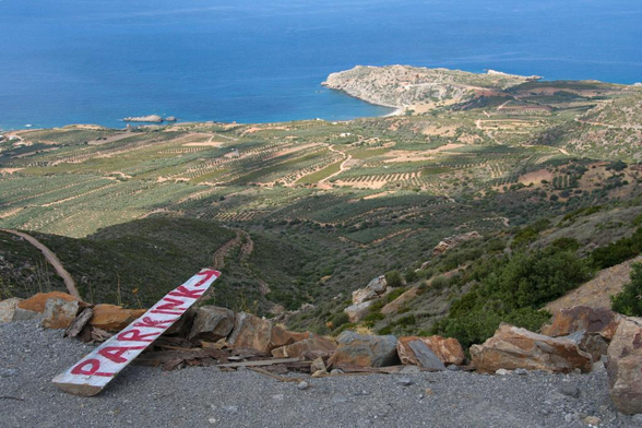 A weathered wooden sign reading “PARKING” rests on rocky ground, overlooking a lush landscape of olive groves and hills that descend to a serene blue sea.