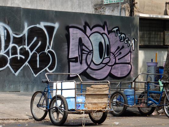 A couple of working style tricycles with two wheels at the front around a metal framed carrying space. Each has a 20litre water bottle in the case. They are haphazardly parked in front of graffiti on a cement wall