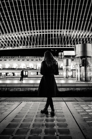 The image is a black and white photograph taken at a train station at night. The main subject is a woman standing on the platform, facing away from the camera. She is wearing a long coat and boots. Her hair is long and wavy. The background features the train station architecture, including a complex, illuminated ceiling structure and a train.

The composition is centered, with the woman positioned in the middle of the frame, drawing the viewer's eye. The architecture forms a backdrop, with the illuminated ceiling creating a dramatic overhead pattern. The train, with its lit windows, adds depth and context to the scene.