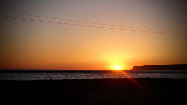 Aus dem Auto bei voller Fahrt auf der Küstenstraße heraus ist der Sonnenuntergang fotografiert. Die Sonne hängt als gelb-oranger Ball direkt am Horizont über dem Meer. Die Klippen der Küste im Hintergrund und der Dünenstrand im Vordergrund sind nur noch dunkle Schatten. Nur auf dem Meer liegt noch ein letzter Silberglanz. Der Himmel mit Farbverlauf in graublau und orange macht sich bereit für die blaue Stunde. 