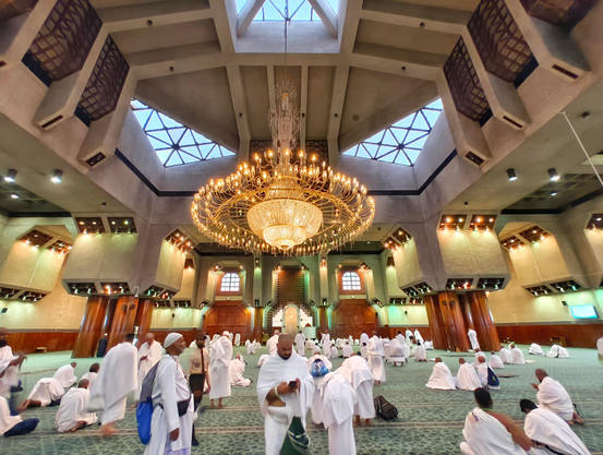 Pilgrims in white attire gather inside a grand mosque, beneath a magnificent golden chandelier. The spacious hall features patterned green carpets, large pillars, and unique skylight windows, creating a serene atmosphere for worship and reflection.