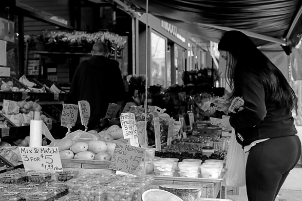 Despite the chill of the fall day, customers shop the fresh produce outside a neighbourhood market. A man in the background has his back turned to the camera. Filling the frame at right, a woman holds a single berry delicately between her fingers.