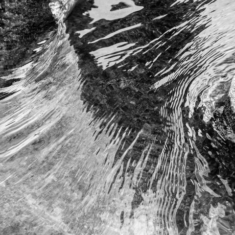 A dramatic black and white photograph captures water in motion, creating dynamic abstract patterns. The image shows flowing water with strong contrast between light and dark areas, featuring streaking patterns and ripples that create diagonal lines across the frame. The water's surface displays texture and movement, with white highlights indicating turbulence or spray against darker areas. The composition is abstract and artistic, emphasizing the fluid, ever-changing nature of water through motion blur and high contrast monochrome treatment.
