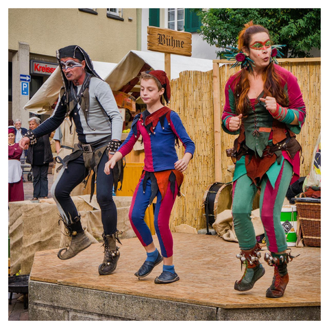 Three performers in vibrant medieval-fantasy costumes are captured mid-jump on a small wooden outdoor stage during a sunny street festival in a German town. The leftmost is an adult man dressed as a pirate or rogue, wearing a black tattered hood, white skull-like face paint around his eyes and mouth, a gray sleeveless jerkin over a light shirt, black pants with a white stripe down one leg, and rugged brown boots adorned with bells and straps, leaping forward with an intense expression. In the center, a young boy in a jester outfit sports a red pointed cap with bells, a blue-and-red split tunic and matching leggings with a black collar, and simple blue shoes, jumping with relaxed arms and a neutral look. On the right, an adult woman as an elf or fairy has long reddish-brown hair in a high ponytail with pink flowers and green antennae-like headpieces, green face paint with red dots and leaf patterns, a green-and-maroon laced corset, turquoise-and-magenta leggings with leaf details, and brown lace-up boots with bells, mid-leap with her right fist raised and a playful puckered-lip expression. The background features a light-colored building with green shutters, a wooden sign reading “Bühne” for stage, a beige canvas tent, woven reed fencing, a large drum, wicker basket, wooden crates, a few onlookers in mixed period and modern clothing, creating a festive and energetic atmosphere.