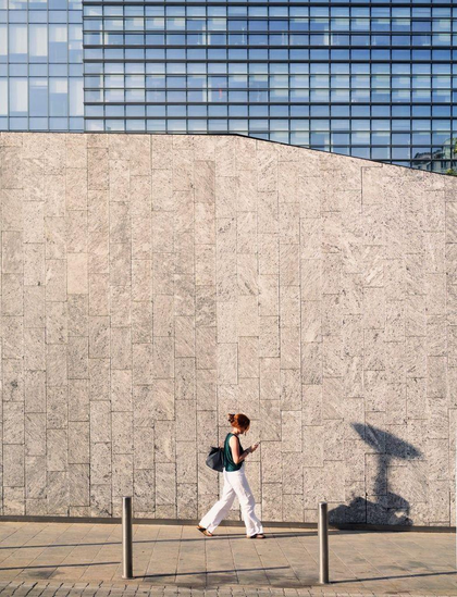 A woman walks along a plain stone wall with a glass building in the background. In front of here, on the wall, is a shadow whose shape doesn't match that of the woman.