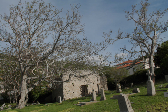 Dieses Bild zeigt einen verlassenen Friedhof mit einer alten, ruinösen Steingebäude im Hintergrund.  Die Bäume sind kahl, was auf die Jahreszeit, möglicherweise Herbst oder Winter, hindeutet. Die Grabsteine sind einfach und alt, was auf eine lange Geschichte des Friedhofs hinweist.  Das Gesamtbild wirkt ruhig und melancholisch, mit einem Hauch von Verfall und dem unausweichlichen Lauf der Zeit. Die Szenerie ruft ein Gefühl der Stille und Reflexion hervor. Im Hintergrund ist ein modernes Haus sichtbar, das einen Kontrast zur antiken Umgebung bildet und die Unterschiede zwischen Gegenwart und Vergangenheit betont.  Das Bild könnte als Metapher für die Vergänglichkeit interpretiert werden, wobei die Ruinen und die verlassenen Gräber den Zerfall repräsentieren, während das moderne Haus die fortdauernde Gegenwart symbolisiert.