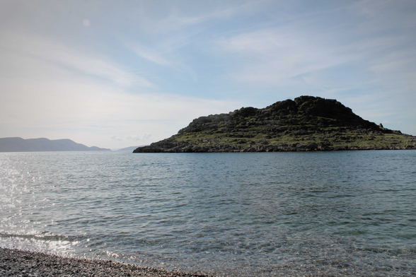 Das Bild zeigt eine ruhige Bucht mit kristallklarem Wasser und einem felsigen Hügel im Hintergrund. Der Himmel ist hellblau mit nur wenigen Wolken, was auf einen ruhigen und sonnigen Tag hindeutet. Das Wasser ist ruhig und reflektiert das Licht, während kleine Wellen sanft an den Kiesstrand schlagen. Die felsige Insel wirkt rau und unberührt, ein Kontrast zur ruhigen Atmosphäre des Wassers. Die Szene vermittelt ein Gefühl von Frieden, Ruhe und Einsamkeit, ein idealer Ort zur Entspannung und zum Genießen der Natur.  Das Bild könnte ein Urlaubsfoto sein, das die Schönheit einer unberührten Landschaft einfängt. Die Farbpalette ist natürlich und beruhigend, mit sanften Blautönen im Wasser und den Grautönen des Felsens. Die Komposition ist einfach, aber effektiv, mit dem Fokus auf der Insel und dem Kontrast zwischen Land und Meer.