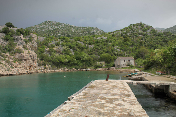 Sanfte Hügel umgeben einen kleinen, ruhigen Hafen, dessen Wasser in einem tiefen, klaren Türkis schimmert. Am linken Rand erhebt sich eine steinerne Klippe, die mit üppiger, grüner Vegetation durchzogen ist. Die unterschiedlichen Grüntöne von Laub und Sträuchern schaffen eine harmonische Kulisse entlang der Felsen, deren raue Textur durch die unterschiedlichen Lichtverhältnisse stark hervorgehoben wird.  Am rechten Bildrand steht ein schlichtes, graues Gebäude, dessen Dach leicht geneigt ist. Die Oberfläche des Hauses scheint von der Zeit gezeichnet und trägt Spuren von Verwitterung. Neben dem Gebäude befindet sich ein kleiner, bewaldeter Bereich, der einen Kontrast zu den kargen Felsen bildet. Auf der rechten Seite sind einige Boote sichtbar, die am Steg liegen und darauf warten, ins Wasser geschoben zu werden.  Der Weg zum Wasser führt über eine breite, graue Steinmauer, die sich in die Bucht hineinzieht. Auf diesem Weg sind einige unregelmäßige Steine zu sehen, die die Struktur unterbrechen und eine natürliche, rustikale Ästhetik erzeugen. Ein rostiger Pfahl ragt am Ende des Stegs in die Höhe, was den Eindruck von einem langen Zeitabschnitt vermittelt, in dem hier Aktivitäten stattfanden.  Im Hintergrund sind die sanften Hügel sichtbar, die mit dichtem Grün bewachsen sind und im oberen Bereich flach auslaufen. Der Himmel ist von grauen Wolken bedeckt, die eine gewisse Dramatik vermitteln und die Lichtverhältnisse im Bild beeinflussen. Das Gesamtbild strahlt eine ruhige und unberührte Schönheit aus, die den Betrachter in die pazifistische Stimmung des Ortes hineinzieht.