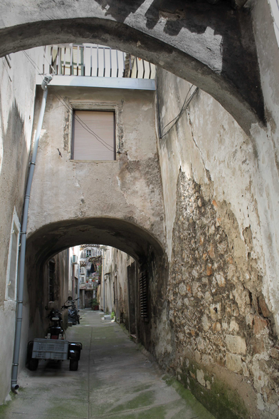 Hier ist eine einfache Beschreibung des Bildes:  Das Bild zeigt eine enge Gasse in einer alten Stadt. Ein Steinbogen überspannt die Gasse und gibt den Blick auf den weiteren Verlauf frei. Die Wände sind alt und verwittert, mit abblätterndem Putz und freiliegendem Stein. Am linken Rand der Gasse steht ein kleiner Wagen. Insgesamt vermittelt das Bild einen Eindruck von Alter, Geschichte und dem Charme einer kleinen, verwinkelten Gasse.