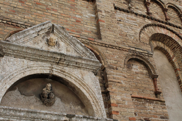 Hier ist eine einfache Beschreibung des Bildinhalts:  Das Bild zeigt einen Ausschnitt eines alten, steinernen Gebäudes. Im Vordergrund ist eine steinerne Fassade mit einem Bogen und einem dreieckigen Giebel darüber zu sehen. An der Wand des Gebäudes befinden sich mehrere dekorative Elemente und architektonische Details wie weitere Bögen. Die Struktur besteht aus roten Backsteinen und Steinen, was ihr ein rustikales und historisches Aussehen verleiht. Das Bild ist aus einer schrägen Perspektive aufgenommen, die Details der Architektur hervorhebt.