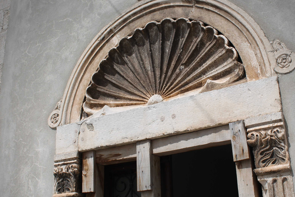 Dieses Bild zeigt einen verwitterten Steintürrahmen mit einem muschelförmigen Ornament über der Tür. Der Rahmen ist aus hellem Stein gefertigt und zeigt Anzeichen von Alterung und Verfall.  Das Muschelmotiv ist detailliert gearbeitet und deutet auf einen möglichen Bezug zur Meereswelt oder zur klassischen Architektur hin.  Die Tür selbst ist mit Holzbrettern provisorisch repariert, was auf einen Zustand des Verfalls oder der Renovierung hinweist. Die Kombination aus verziertem Stein und provisorischer Holzreparatur erzeugt einen interessanten Kontrast zwischen alter Pracht und aktuellem Zustand, und unterstreicht die Geschichte des Bauwerks. Das Bild wirkt insgesamt ruhig und evoziert ein Gefühl von Geschichte und Vergänglichkeit.