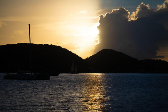 Sunset over the harbor in Charlotte Amalie, St. Thomas. The sun dips behind dark hills, sending a golden reflection across the calm water. A sailboat and catamaran are silhouetted in the foreground beneath a towering cloud glowing with light.
