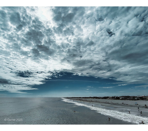 Expansive beach scene with partially cloudy sky, gentle waves, and scattered beachgoers. The mood is serene and calm.
