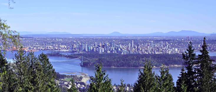 A major city's central business district in the midground, framed by conifers, in the foreground, an inlet and a small forest just in front of the city, and a vast expanse of lower-density city into the background with some secondary centres in the distance. There are mountains across the far background.