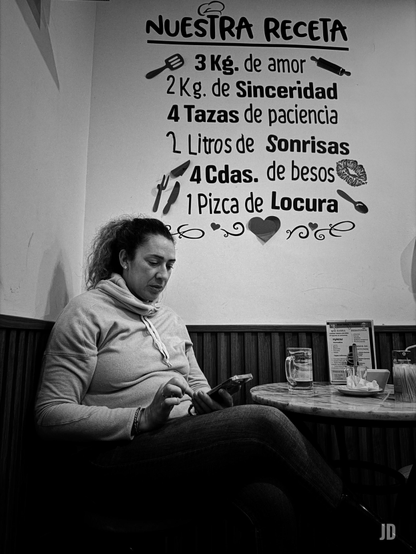 Fotografía en blanco y negro de una mujer sentada en un café o sala, absorta en su teléfono móvil. La mujer, de mediana edad y con el pelo rizado recogido, viste una sudadera con capucha y jeans, y su mirada está fija en la pantalla del dispositivo que sostiene con ambas manos. Detrás de ella, en la pared clara, hay un gran texto decorativo que simula una receta de cocina, titulada "NUESTRA RECETA". La lista de 'ingredientes' incluye elementos como "3 Kg. de amor", "2 Kg. de Sinceridad", "4 Tazas de paciencia", "2 Litros de Sonrisas", "4 Cdas. de besos" y "1 Pizca de Locura", acompañados de iconos relacionados con la cocina (espátula, rodillo, cuchillo, tenedor) y símbolos de afecto como labios y corazones. A la derecha de la mujer, sobre una mesa redonda de madera, se aprecian un vaso con bebida, un menú y una pequeña pila de platos. La parte inferior de la pared está cubierta con paneles de madera.