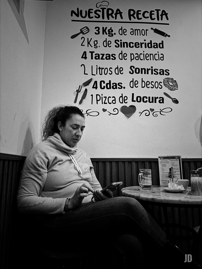 Fotografía en blanco y negro de una mujer sentada en un café o sala, absorta en su teléfono móvil. La mujer, de mediana edad y con el pelo rizado recogido, viste una sudadera con capucha y jeans, y su mirada está fija en la pantalla del dispositivo que sostiene con ambas manos. Detrás de ella, en la pared clara, hay un gran texto decorativo que simula una receta de cocina, titulada "NUESTRA RECETA". La lista de 'ingredientes' incluye elementos como "3 Kg. de amor", "2 Kg. de Sinceridad", "4 Tazas de paciencia", "2 Litros de Sonrisas", "4 Cdas. de besos" y "1 Pizca de Locura", acompañados de iconos relacionados con la cocina (espátula, rodillo, cuchillo, tenedor) y símbolos de afecto como labios y corazones. A la derecha de la mujer, sobre una mesa redonda de madera, se aprecian un vaso con bebida, un menú y una pequeña pila de platos. La parte inferior de la pared está cubierta con paneles de madera.