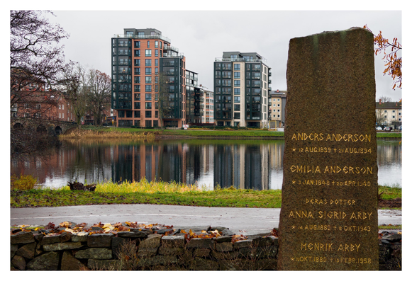 En liggande bild på en gravsten från 1800-talet. Den står vid en mur och på andra sidan älven står två nybyggda flerfamiljshus.