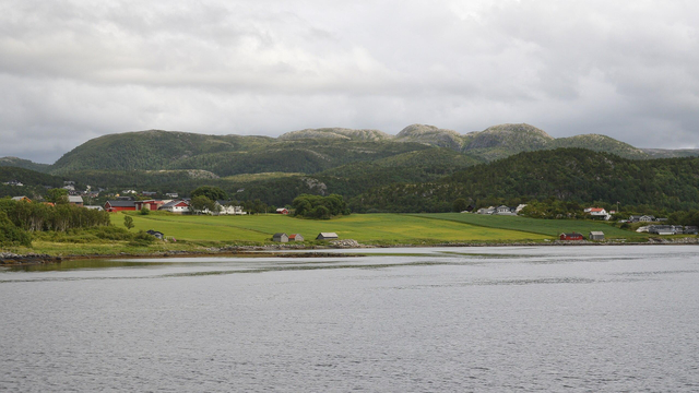 A photo of farm fields on a shore leading into forests with low mountains in the distance. The sky is cloudy.