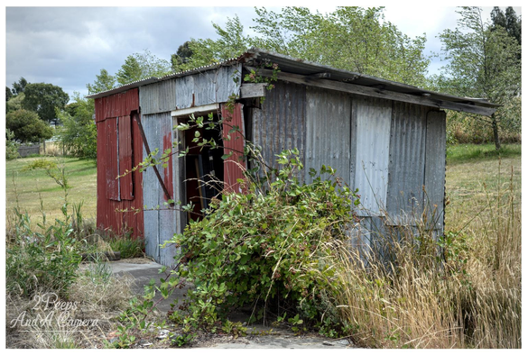 A photo of a small, dilapidated shed made of corrugated iron. The shed is surrounded by overgrown grasses and thick brambles that partially obscure the doorway.

The front of the shed is a patchwork of rusted red and oxidized grey metal, with a lean to roof. The surrounding field is dry and grassy, with trees visible in the distance under a cloudy sky.