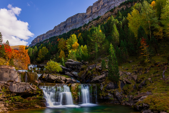 Español: cascada de agua cristalina entre rocas y vegetación otoñal, rodeada de árboles verdes, amarillos y anaranjados, con una montaña rocosa al fondo bajo un cielo azul.

English: A clear waterfall flowing over rocks amid autumn vegetation, surrounded by green, yellow, and orange trees, with a rocky mountain in the background under a blue sky.