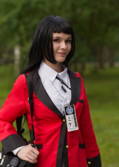 The image captures a person outdoors, standing amidst what appears to be a park or garden. The individual is dressed in an attention-grabbing red coat with black accents and details. A badge hangs prominently from the neck on a lanyard, indicating some form of identification or affiliation. In their right hand, they hold what looks like a small purse or bag. The person's hair is styled in straight bangs that fall over the forehead and are cut to shoulder length at the back.

The background is softly focused but shows an expanse of greenery with trees and possibly other park visitors or structures slightly obscured by the shallow depth of field used for the photograph, which places emphasis on the subject. The lighting suggests it could be a bright day due to the overall brightness and absence of harsh shadows cast by the person's figure.

The person's expression is neutral to slightly smiling, suggesting a posed photograph rather than candidly captured in mid-action or conversation.
