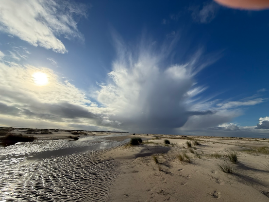 Hinter flachen Sanddünen türmt sich eine von Luftturbulenzen ausgegrenzte Regenwolke in der Mitte des Bildes auf. Rechts von ihr ist der Himmel kräftig blau, während links Du Sonne durch dünne Wolken scheint und die Wolken von hinten beleuchtet.