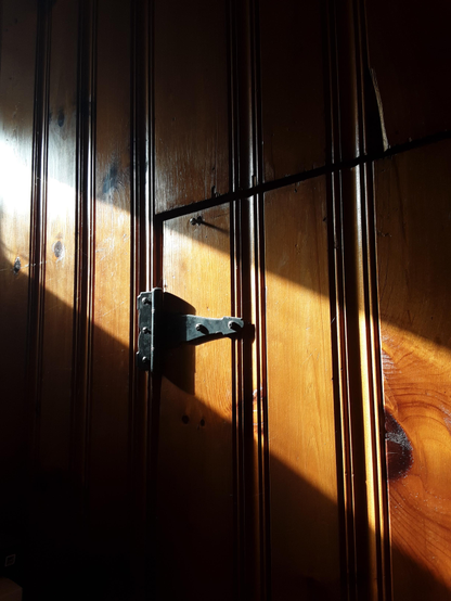 Sunlight diagonally cutting across an old-fashioned wood panelling door with black wrought-iron hinge.