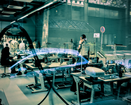 Blick durch das Fenster in einen Raum, in dem Näh- und Bügeltische stehen. Eine schwarzgekleidete Frau mit Hijab sitzt an einem der Tische und arbeitet noch. An einem anderen Tisch steht ein Mann in Feinripp-Unterhemd mit dem Rücken zum Fenster. 
Die Atmosphäre in dem hohen Raum ist kühl, aber das Licht lässt die Hitze der Bügeleisen in dem Raum erahnen. 