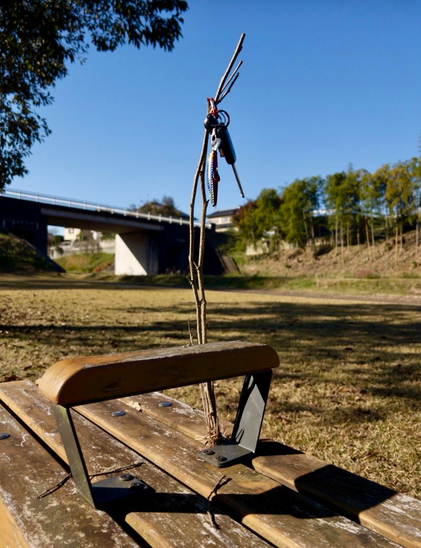 A small wooden bench features a metal support with a stick stuck in it. Someone’s lost or forgotten keys are hanging from the top of the stick. The background shows a grassy area and a bridge under a clear blue sky.
