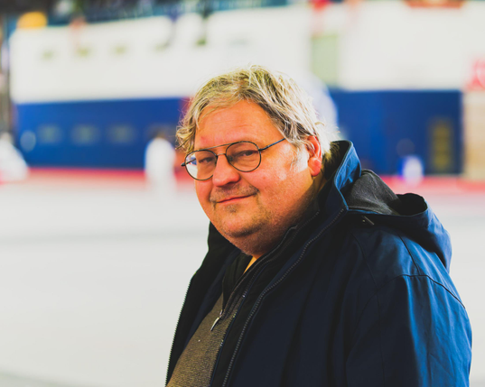 Middle age man is near the center of the frame with a slight smile, round glasses, grey hair and a blue jacket.  Behind him is a building inside a building striped blue on the bottom and white on top.