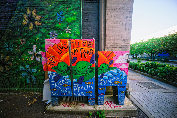 To the right is a plaza with brick walkways and green trees and shrubs,  right in the middle some electrical boxes have a mural painted on them showing three peas rowing a bow underneath a red sun.