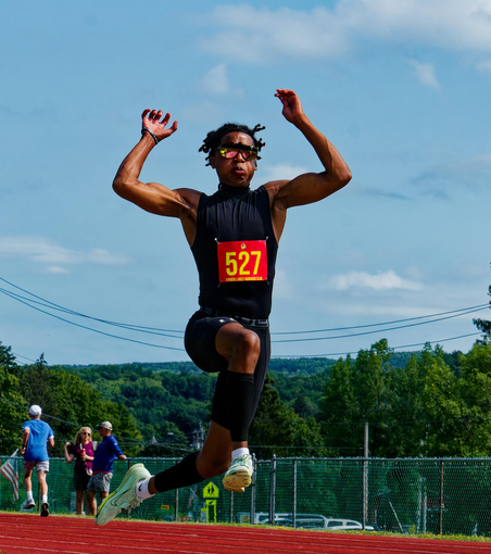 Black man with black clothes and white shoes looks suspended in the air over a red track and with a fence, road and lots of trees behind him