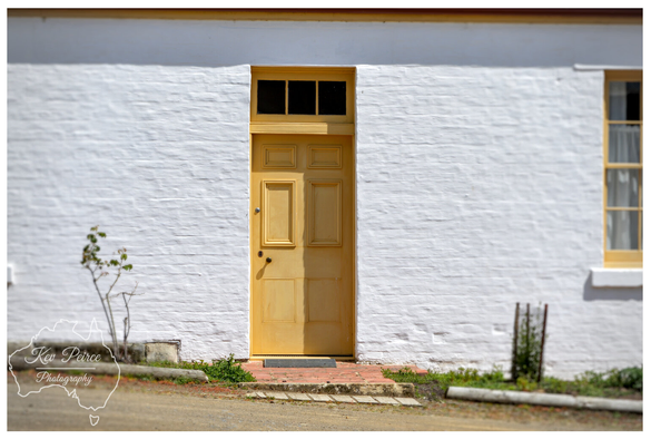 A historic white painted brick wall with a single, brightly painted mustard yellow wooden door in the center.

The door has a rectangular window transom above it, also painted yellow. A small step leads up to the door from a dirt path in the foreground.