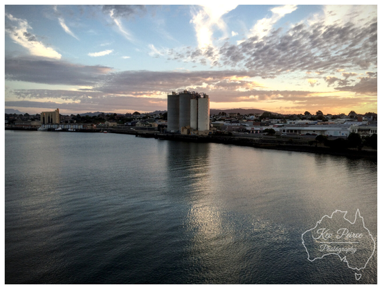 A wide angle photo of the Devonport waterfront in Tasmania at sunset.

The calm waters of the Mersey River reflect the sky, which features high, streaky clouds illuminated by the setting sun's warm glow on the right.

In the middle ground are three prominent, grey cylindrical grain silos and other port buildings lining the shore, with the town's residential area visible in the distance. 

The image is signed Kev Peirce.