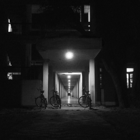 A black-and-white nighttime photograph showing a corridor illuminated by a series of ceiling lights that create a vanishing point effect. Bicycles are parked in the foreground under the first light, while the surrounding area is dimly lit. The structure is a part of hostels.