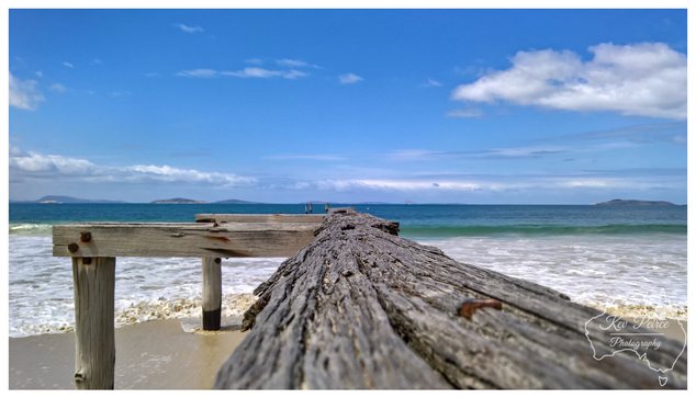 A wide angle, low perspective photograph of a heavily weathered wooden pier or jetty leading out into the ocean from a sandy beach.

The aged wood shows deep grooves and texture, dominating the foreground as it angles sharply from the lower left towards the center.

Gentle waves wash onto the wet sand on either side. In the background, the sea is a deep blue-green, meeting a clear blue sky dotted with white clouds.

Several small, rocky islands are visible on the distant horizon.