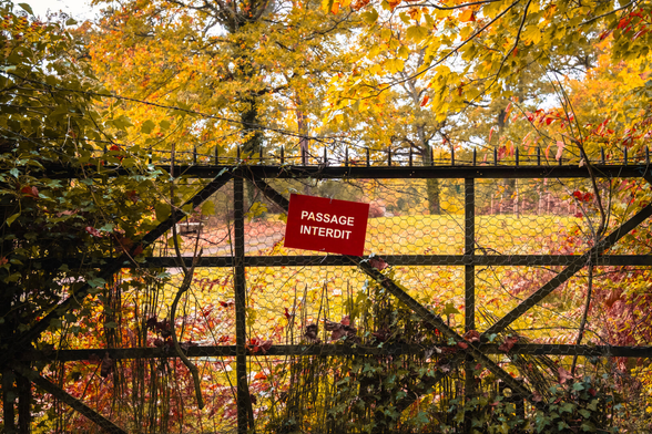 A fence in bad shape with barbwire and a sign of forbidden access before a gorgeous garden in autumn