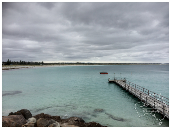 A wide angle landscape photo of a calm turquoise coloured ocean with a long wooden jetty extending from the right foreground.

The jetty leads out toward a small red object floating in the water. The beach runs along the left side of the frame, backed by low coastal scrub and houses under a heavily clouded, grey sky.