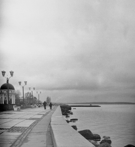 In the black-and-white photo on the left, the embankment of Lake Onega stretches into the distance. In the background, you can see a rotunda with a spire, lanterns, and people's figures. On the right, there is a calm lake with a cloudy sky.