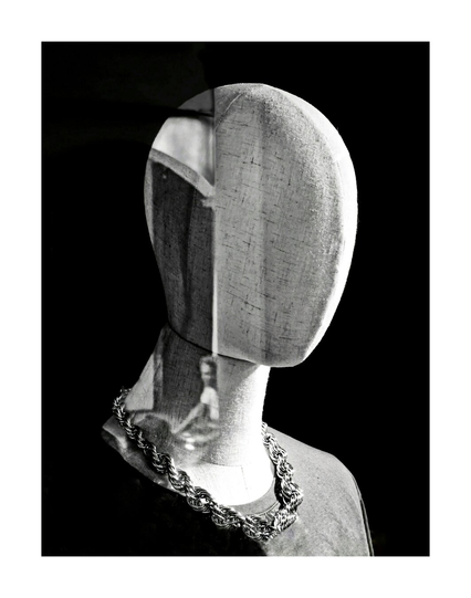 This is a black and white photograph of a faceless mannequin from the shoulders up. It has a textured, fabric-covered head and neck. The mannequin is wearing a shirt and a thick, chunky chain necklace. The background is solid black. The head and neck have a reflection on them from the street - a slice of porticoe with a person riding away on a bicycle.