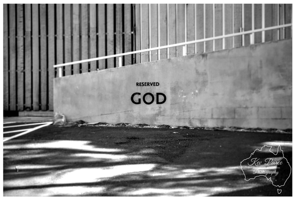 A black and white photograph of an urban scene featuring a concrete retaining wall with a subtle slope.

The text "RESERVED" is written above the large, bold word "GOD" painted or stenciled in black on the smooth surface of the wall, marking a parking spot.

A white metal railing runs along the top of the wall. In the background, there is a vertically corrugated metal fence or structure, while the foreground asphalt shows strong shadows cast across it.