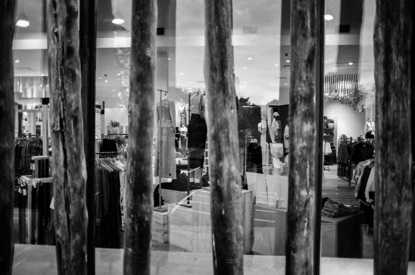 A black and white photo of a retail store taken through the window.  There are vertical wooden battens used as decorations next to the window.  Various clothing items are hanging on racks inside the store.