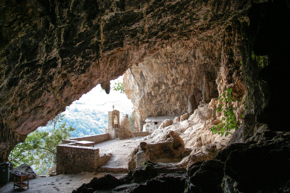 An interior view of a cave featuring a stone altar with a cross, a table, and rough stone surfaces. Natural light enters from an opening.