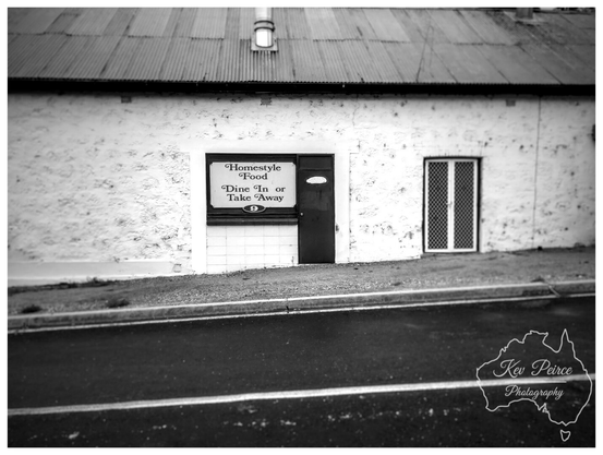 A black and white photograph signed by Kev Peirce showing a modest building with a textured white stone or rendered wall and a corrugated iron roof.

The image is taken from across a street with an asphalt road and a slight grassy embankment. In the center is a simple shopfront with a sign in the window that reads "Homestyle Food Dine In or Take Away."

There is a dark wooden door to the right of the window and a screened white door further right. The image has a vintage or artistic feel due to the black and white conversion and shallow depth of field.