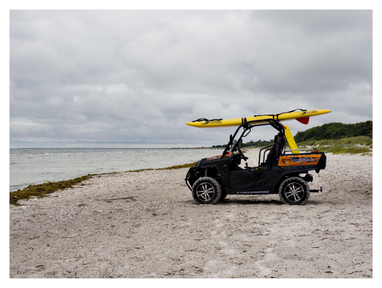 En liggande bild på ett svart, öppet fordon på en sandstrand. På taket sitter en gul surfingbräda fastsurrad. Till vänster är havet och längre bort till höger gräs och skog. Himmelen är full av moln med lite struktur.