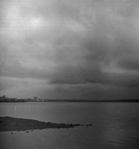The black-and-white photo shows the surface of Lake Onega and the gloomy autumn sky with clouds. It is late evening. In the distance, you can see the shore and the lights of the houses on it. The shoreline is also visible in the foreground.
