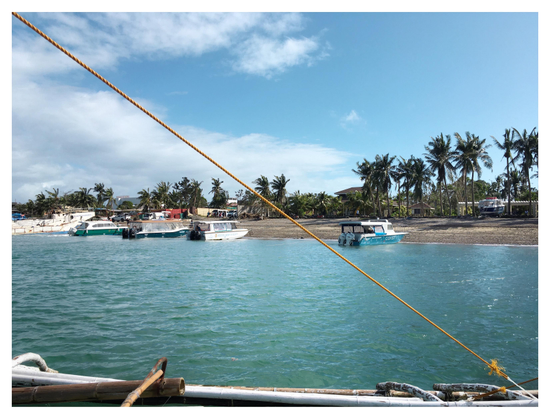 Photo of an island beach as seen from an approaching boat: coconut trees, blue sky with white clouds, boats anchored near the shore. A taut yellow rope, fastened to a boat outrigger at bottom frame, stretches from the upper left corner to the lower right corner of the photo and diagonally bisects the view.