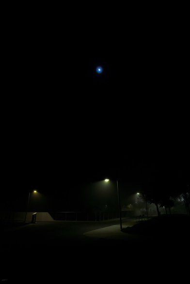 Das Bild zeigt eine nächtliche Szene mit einer geheimnisvollen, leicht nebligen Atmosphäre. Oben im Bild leuchtet der Mond als bläulicher Lichtpunkt mit einem Halo am dunklen Himmel. Unten wird ein Weg von mehreren Straßenlaternen beleuchtet, die ein sanftes, gelbliches Licht auf den Boden werfen. Die Umgebung wirkt ruhig und fast mystisch durch die Kombination aus Dunkelheit, Mondlicht und künstlicher Beleuchtung.