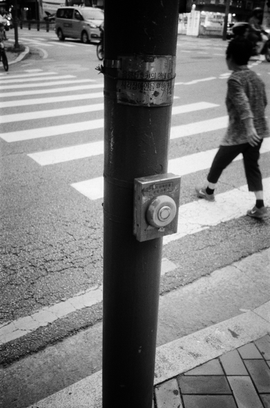 '시각 장애인용 음향 신호기' 버튼이 신호등 기둥에 달려있다. 뒤쪽으로 횡단보도를 건너는 사람이 보인다.