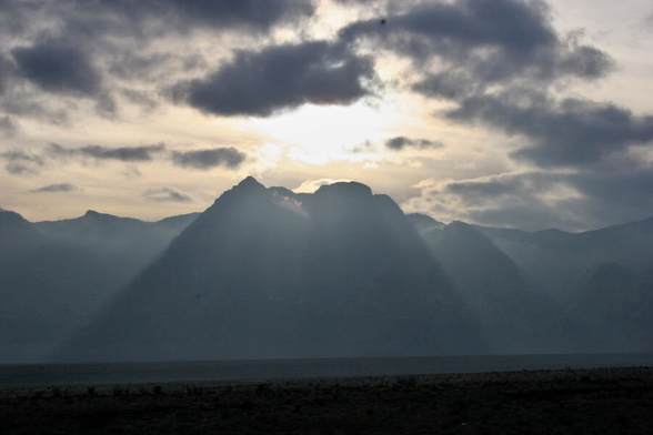 The sun sets behind desert mountains and clouds, casting rays down the mountainside